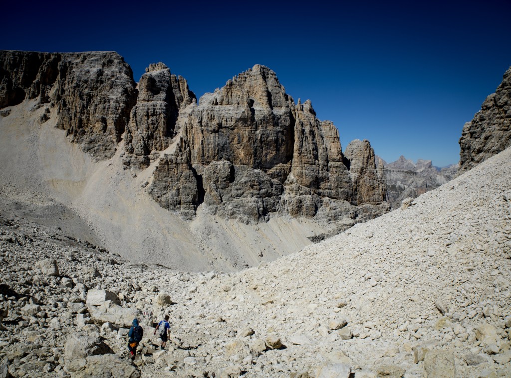 Two hikers walking on a rocky trail in a mountainous landscape with towering cliffs and a clear blue sky.